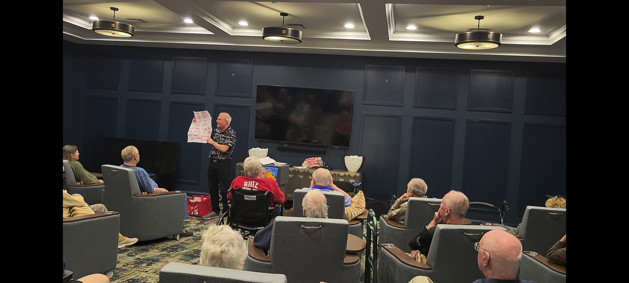 498174401_1231438995444934_2769874638786477557_njpg | Rick Deezie | Rick Deezie Older man stands at front of a blue-paneled room, holding a handwritten poster as a seated audience watches a presentation.