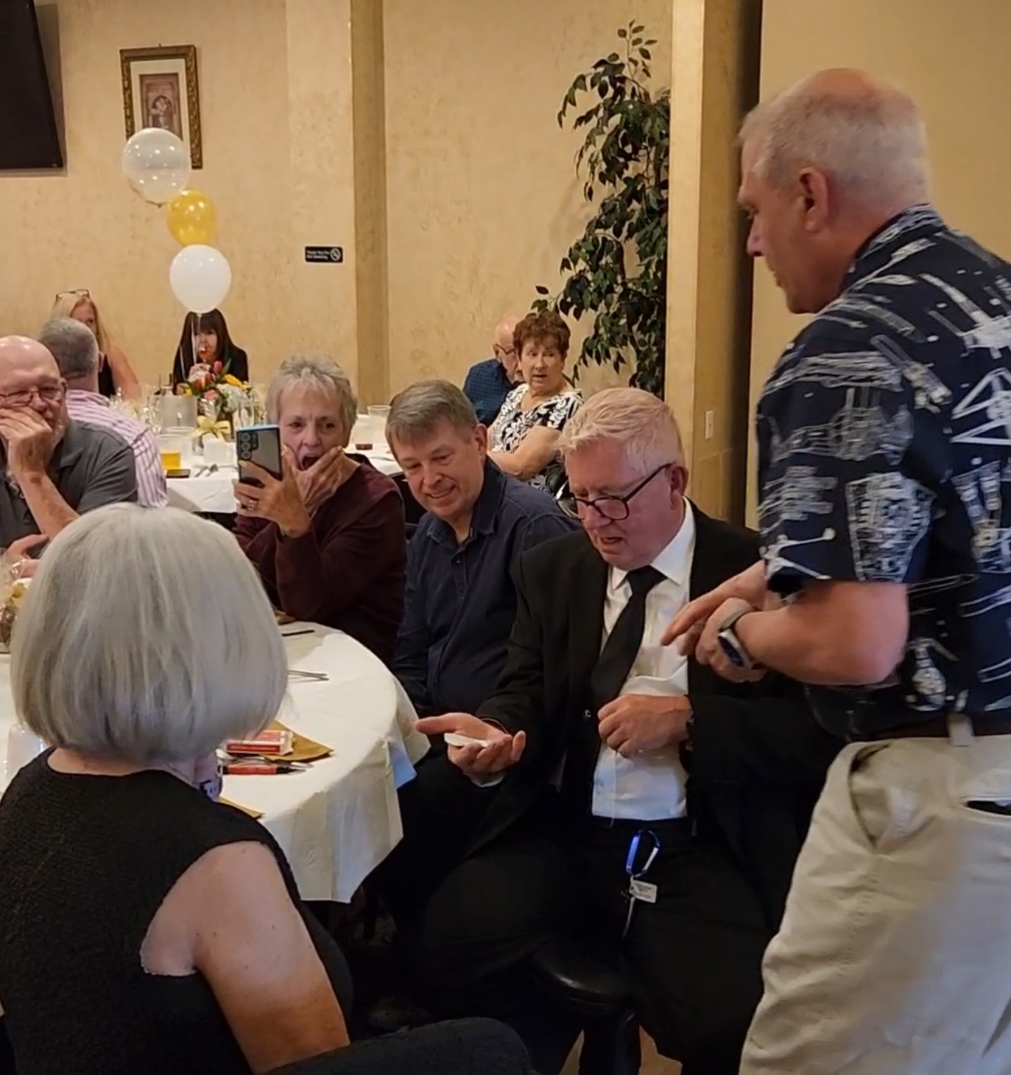 Screenshot_20250515_082455_Video-Playerjpg | Rick Deezie | Rick Deezie Group of people seated at a banquet table listening as a man in a patterned shirt speaks to a guest in a suit and tie at a formal event.