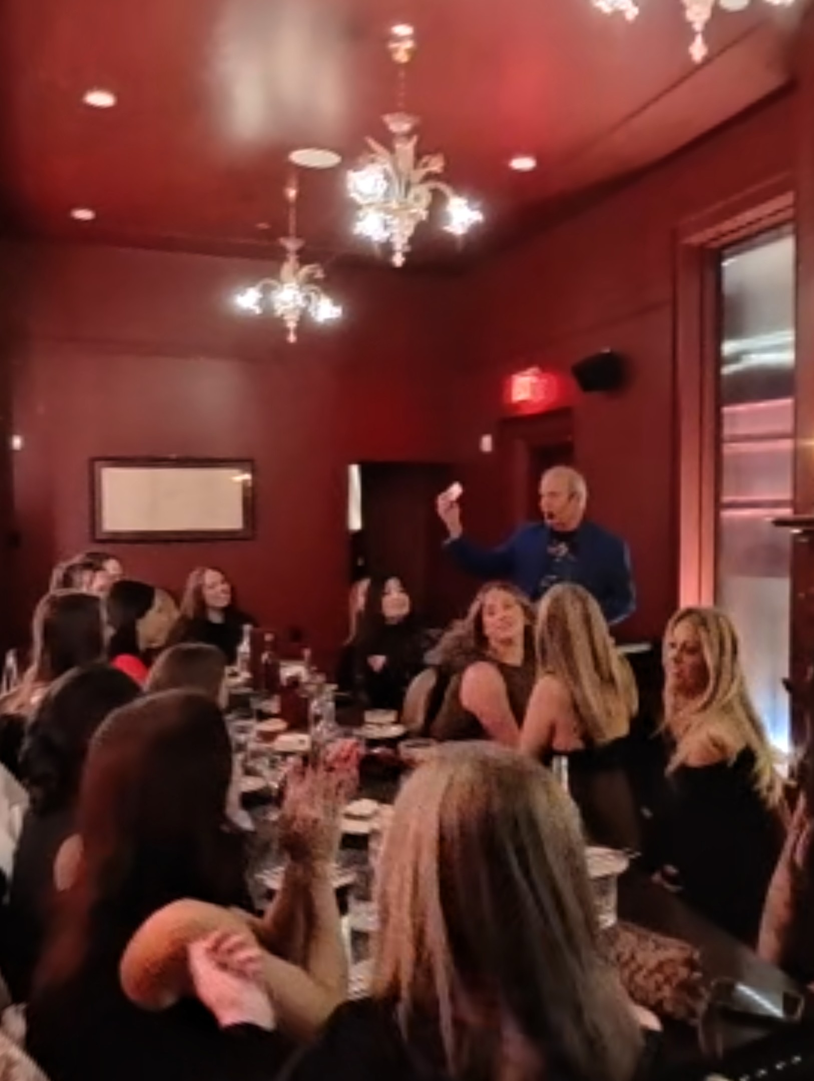 Tatianna-Party-Verticaljpg | Rick Deezie | Rick Deezie Speaker in a blue jacket addressing guests at round banquet tables in a red-walled room with chandeliers.
