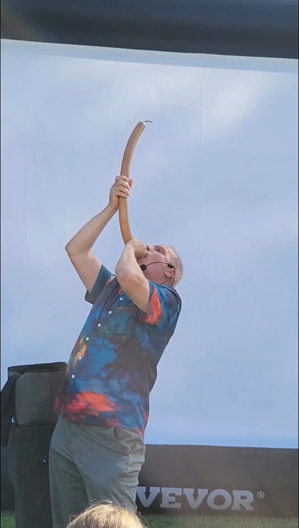 image-29jpg | Rick Deezie | Rick Deezie Man in a tie-dye shirt blows into a long curved horn on an outdoor stage, wearing a headset microphone.
